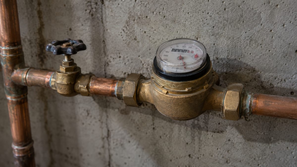 A brass residential water meter connected to copper piping beside a basement wall.